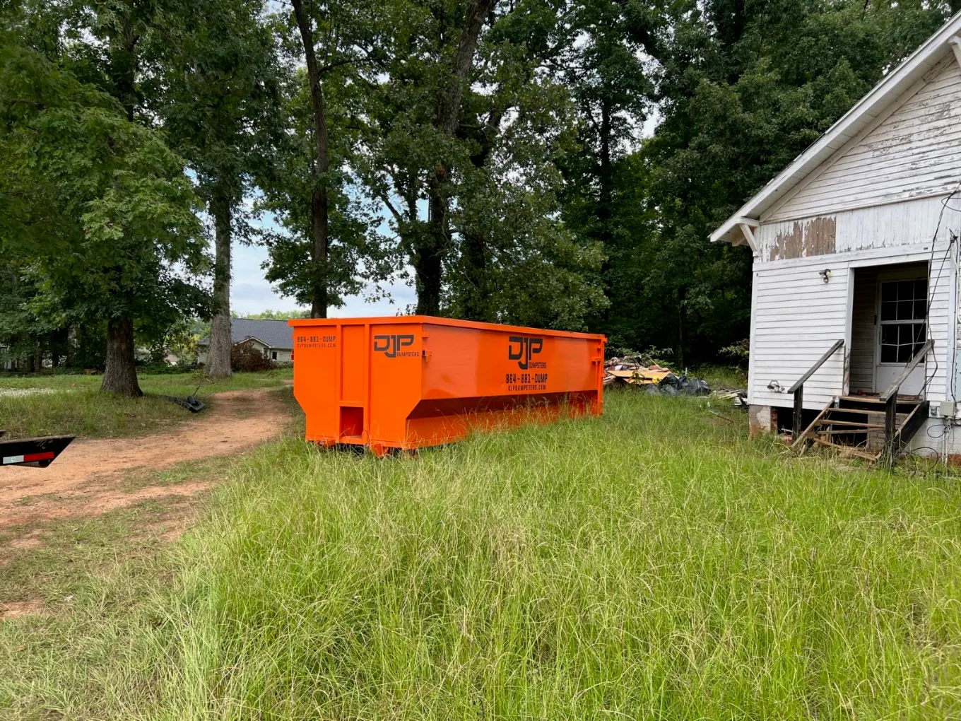 A large orange DJP dumpster beside an old white farmhouse with a debris pile alongside