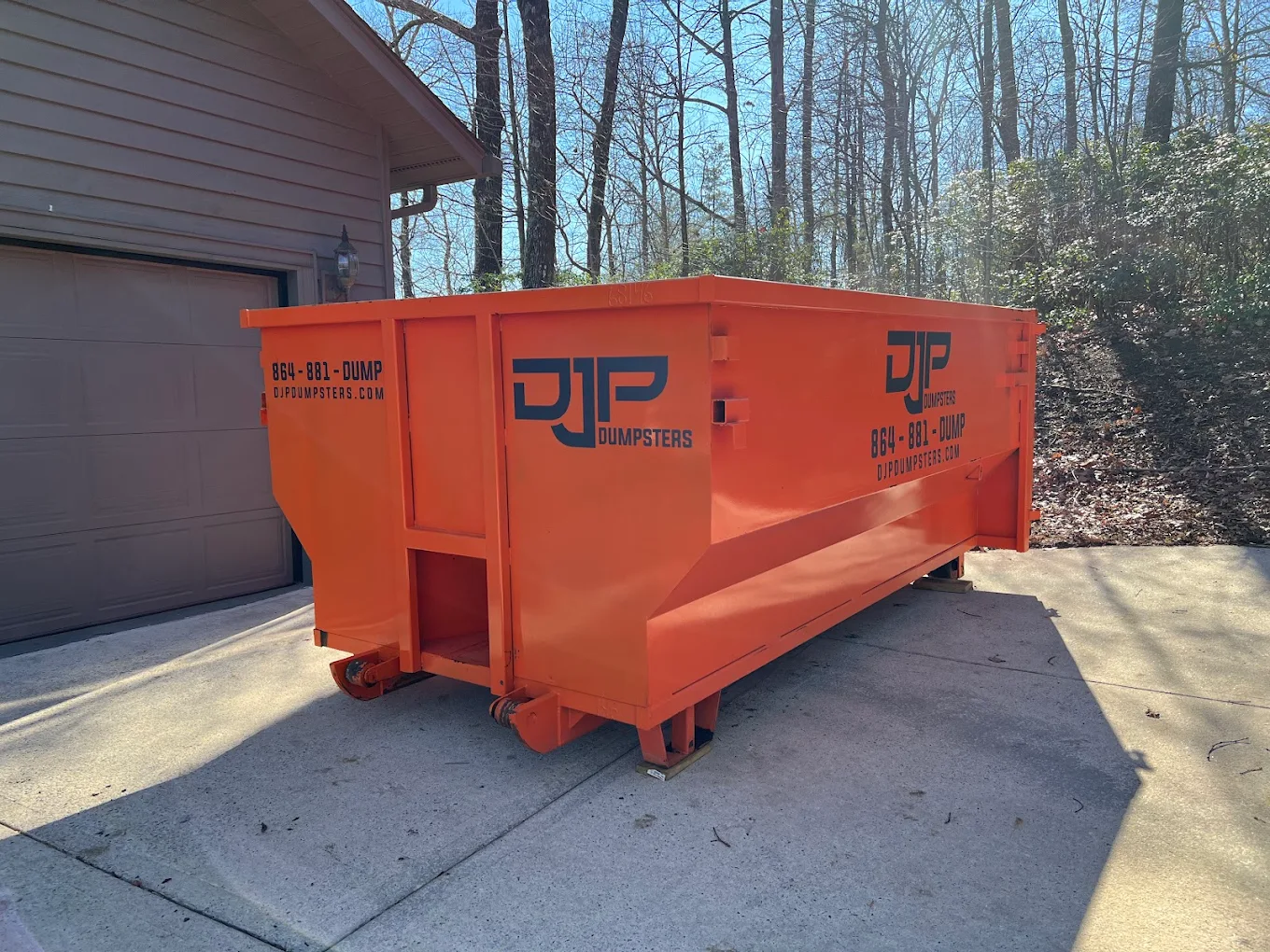 An orange DJP dumpster set on a residential concrete driveway, resting on protection boards