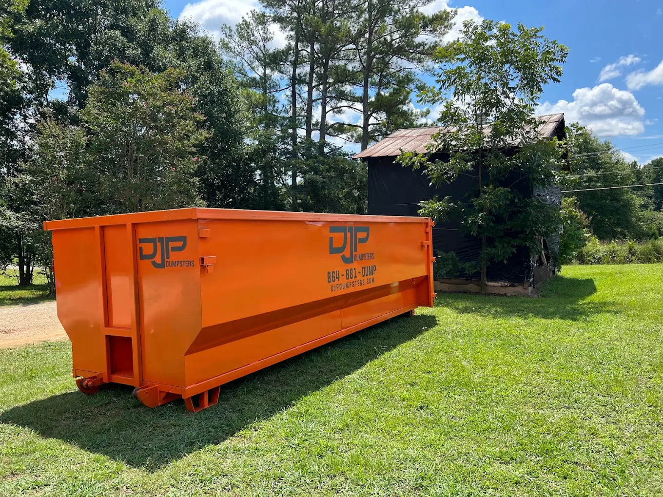 A large orange DJP dumpster on a lawn in front of an old tin-roof barn
