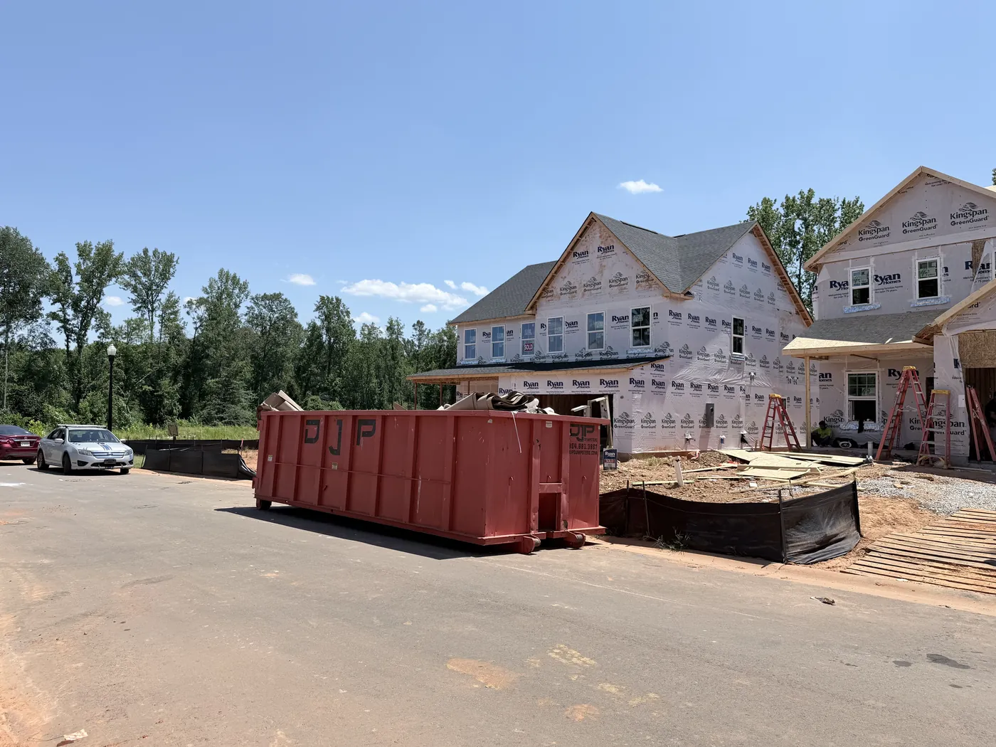 An orange DJP dumpster on a new-construction street with a telehandler and roll-off truck behind it