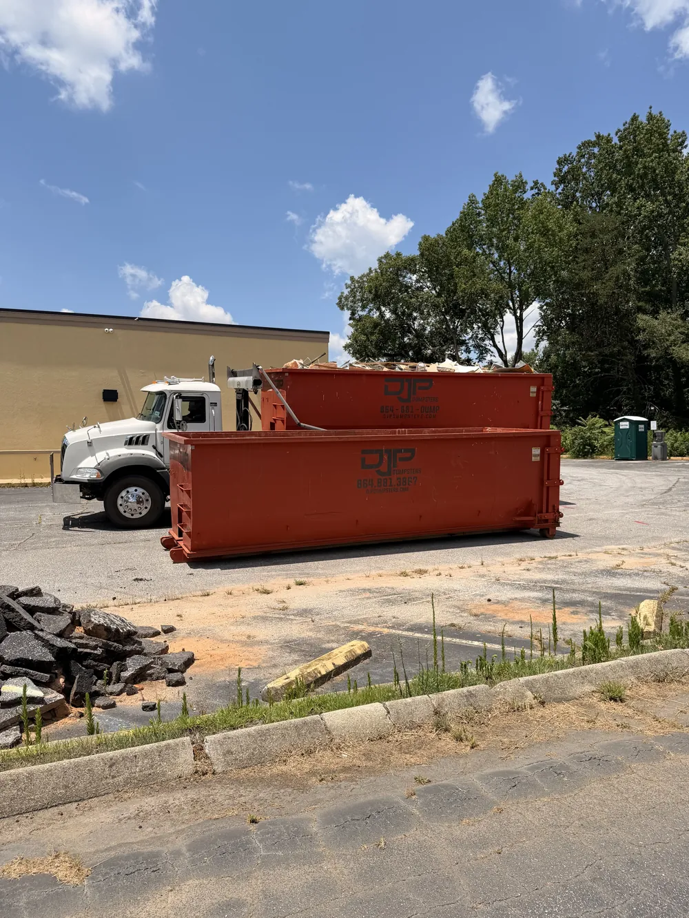Two DJP roll-off dumpsters and a truck on a commercial lot, one loaded with broken asphalt and demolition debris