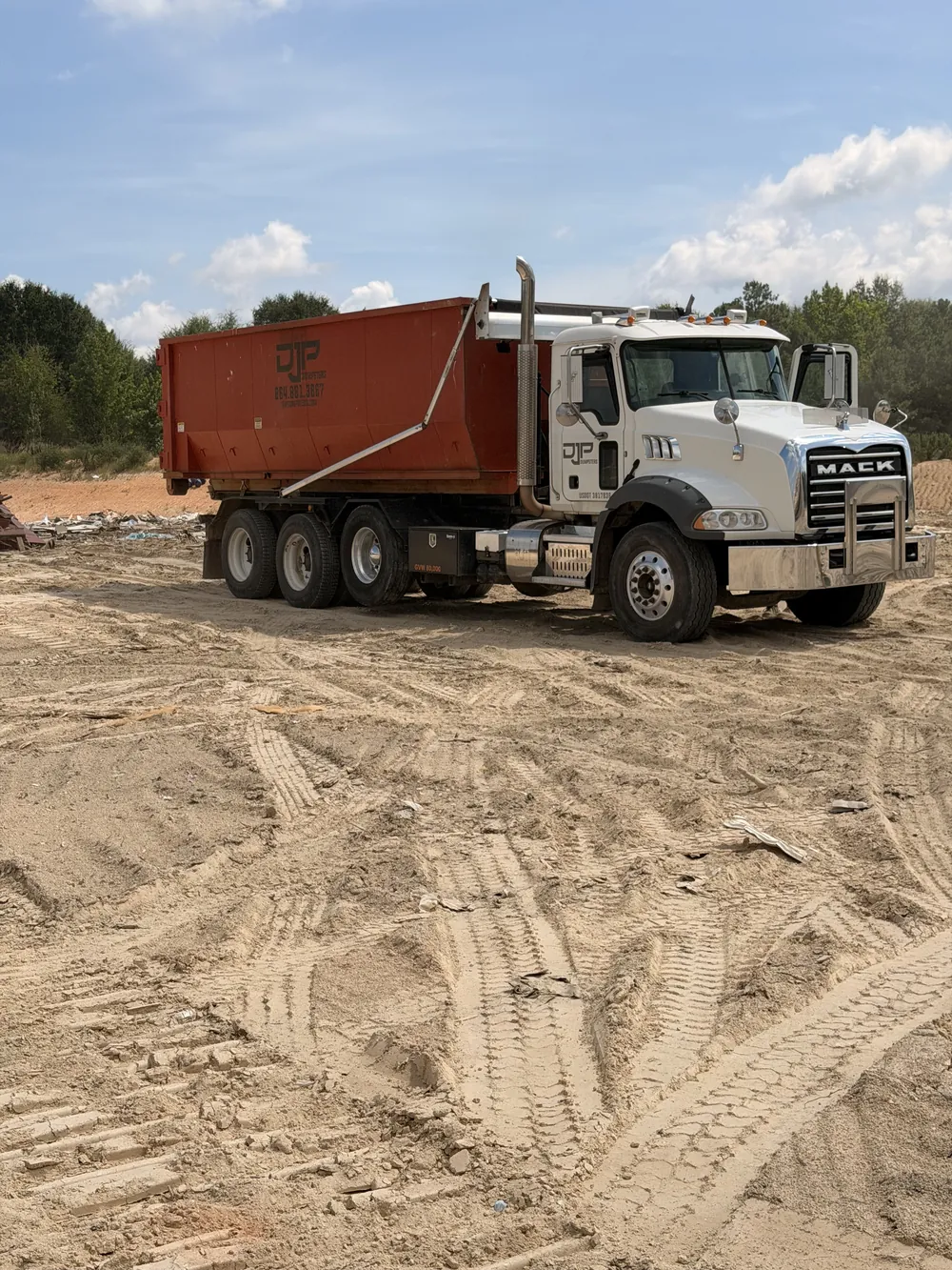 The DJP Mack roll-off truck with a dumpster loaded, parked on a cleared dirt site